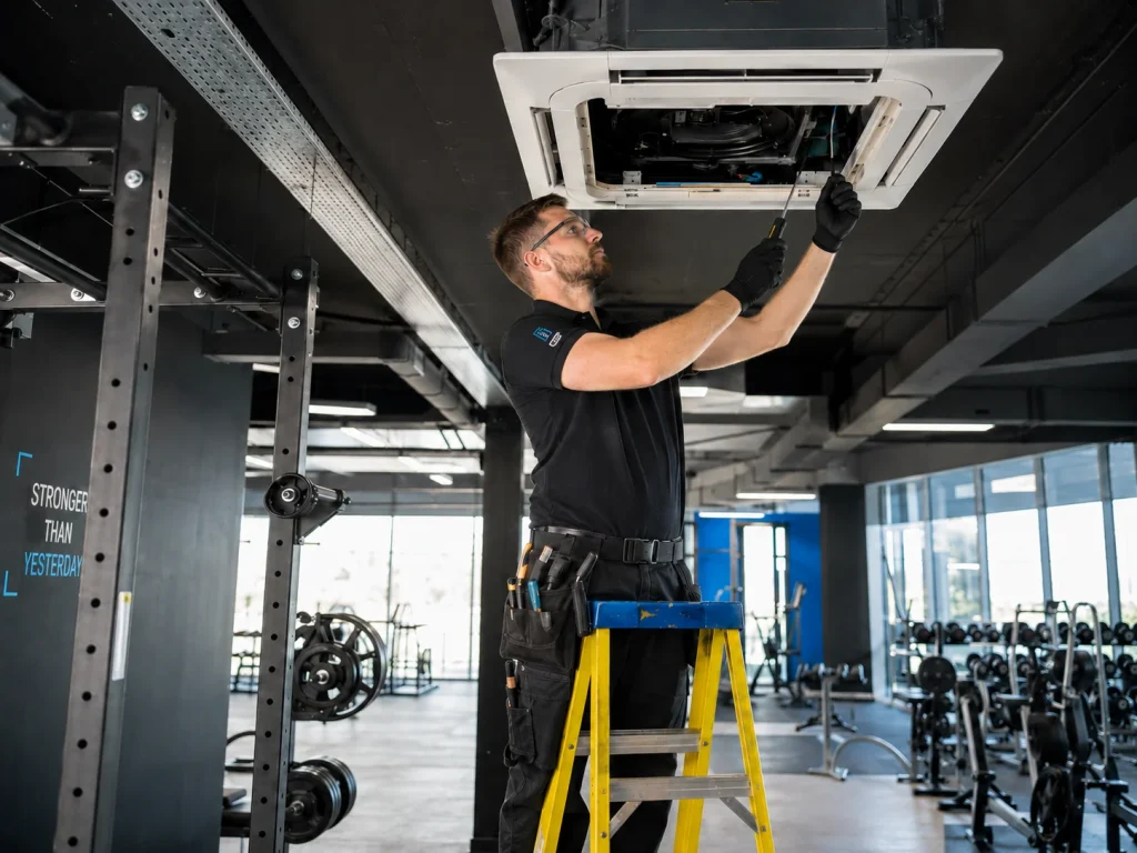 Engineer carrying out gym air conditioning maintenance on a ceiling cassette system in an empty fitness centre.
